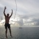 man jumping into water off rope swing while on charter with luxury catamaran in view